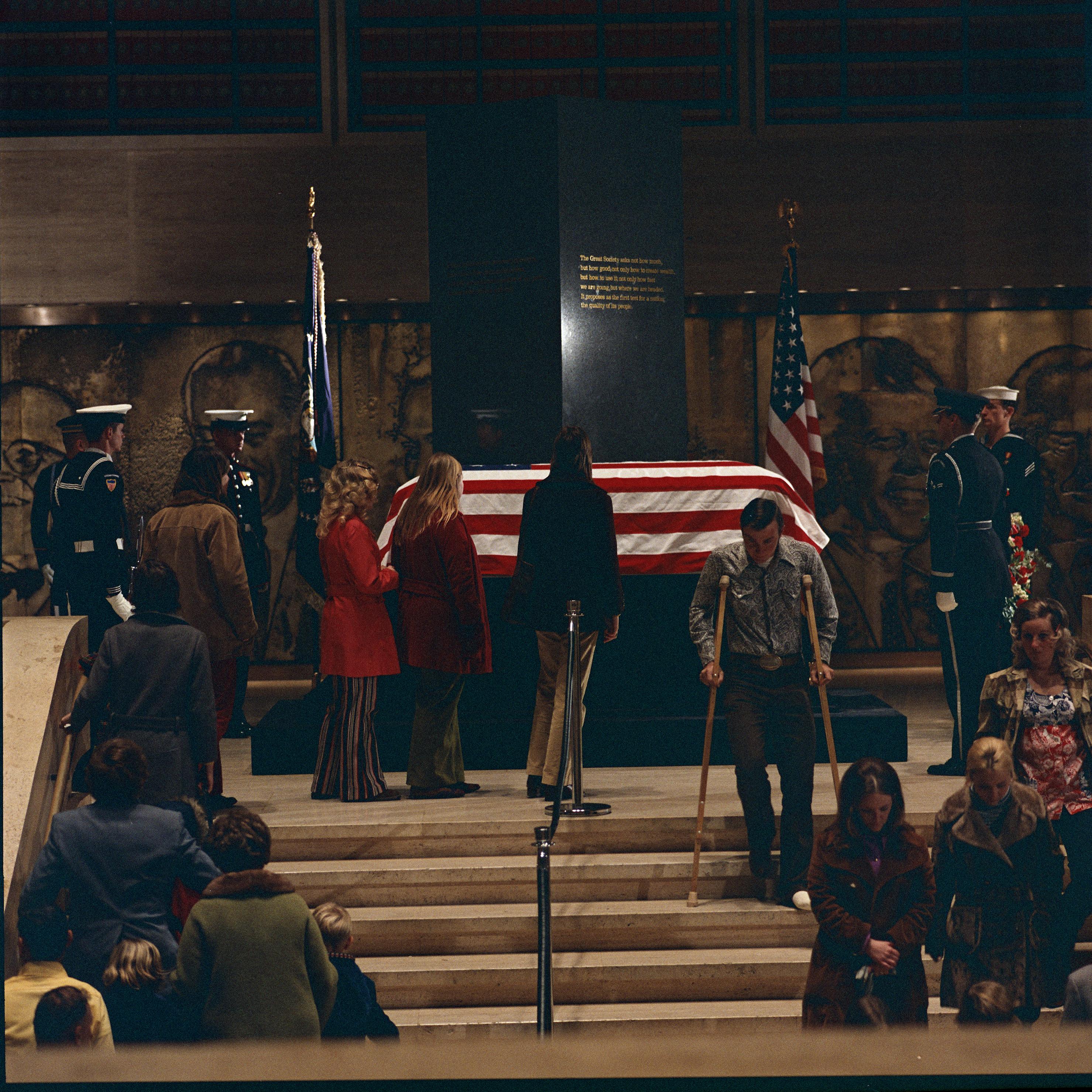 The president's casket, draped in an American flag, lies across the center of the frame, in front of an obelisk. Servicemen stand to the sides, while the public pay their respects. A man with crutches walks down the steps.